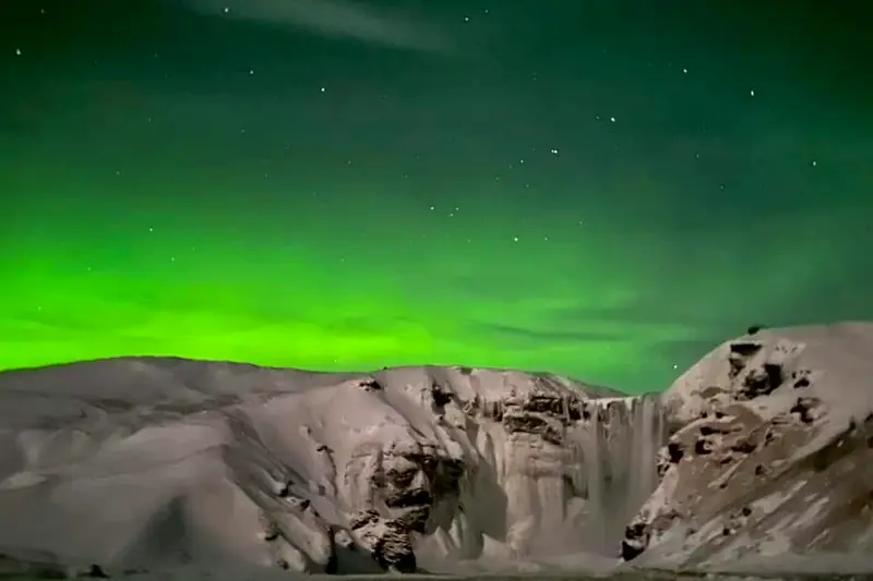 Aurora boreal verde sobre una cascada congelada y montañas nevadas