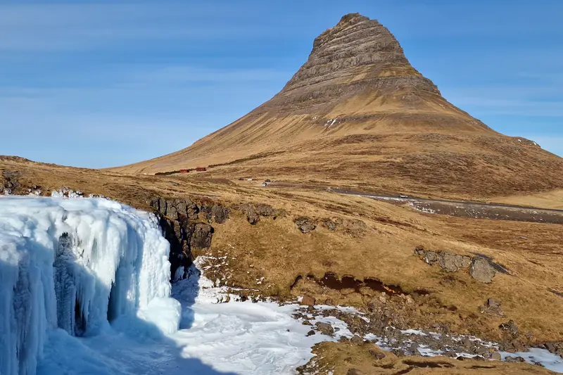 Montaña Kirkjufell bajo cielo azul con la cascada congelada en primer plano