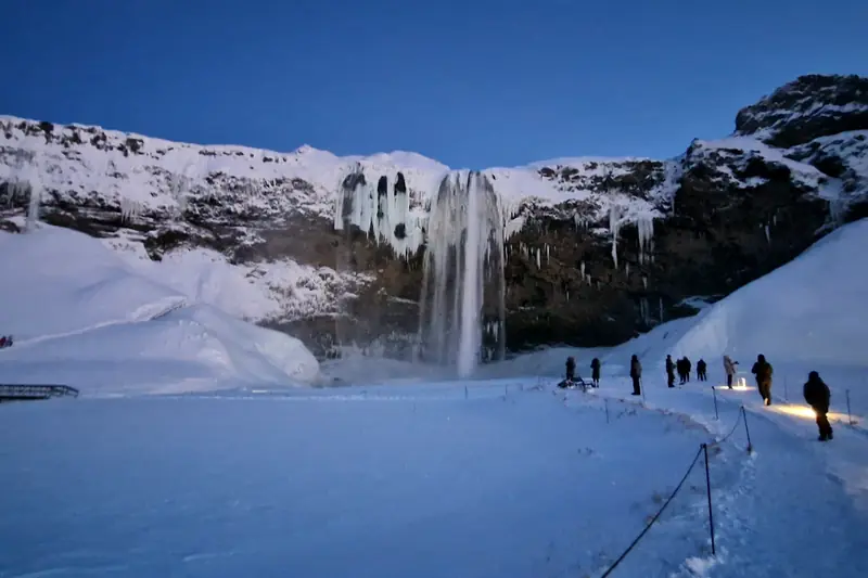Cascada nevada al anochecer con un sendero iluminado y viajeros caminando