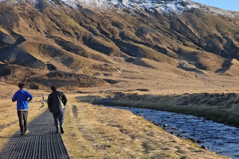 Dos viajeros caminando por un sendero junto a un río entre montañas en otoño